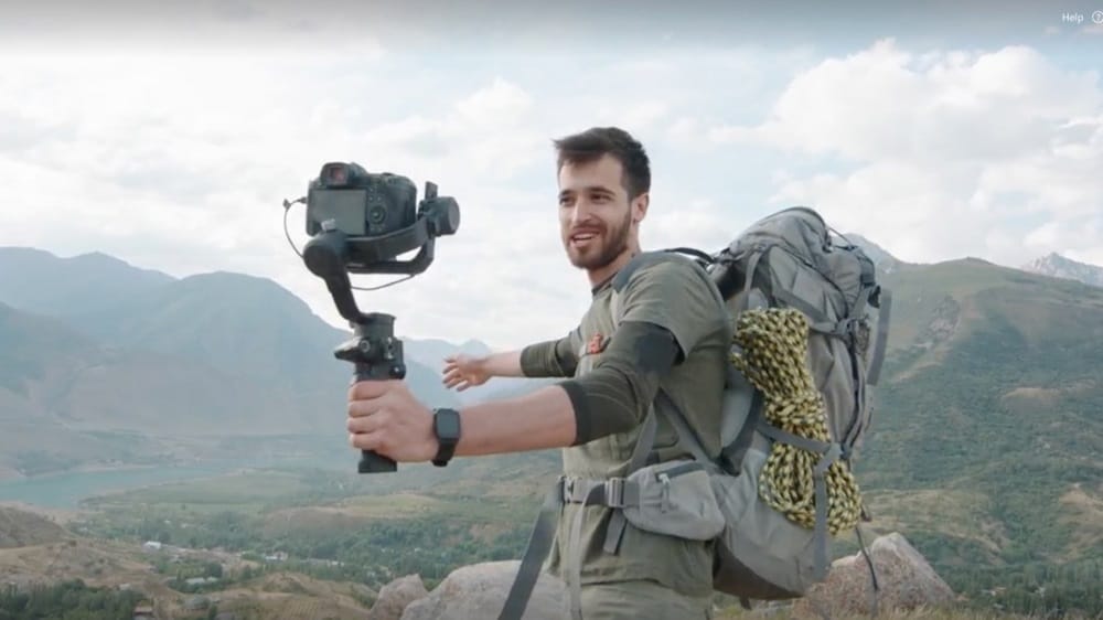 Travel vlogger filming with a DSLR camera on a stabilizer while hiking in the mountains with a backpack and climbing rope.