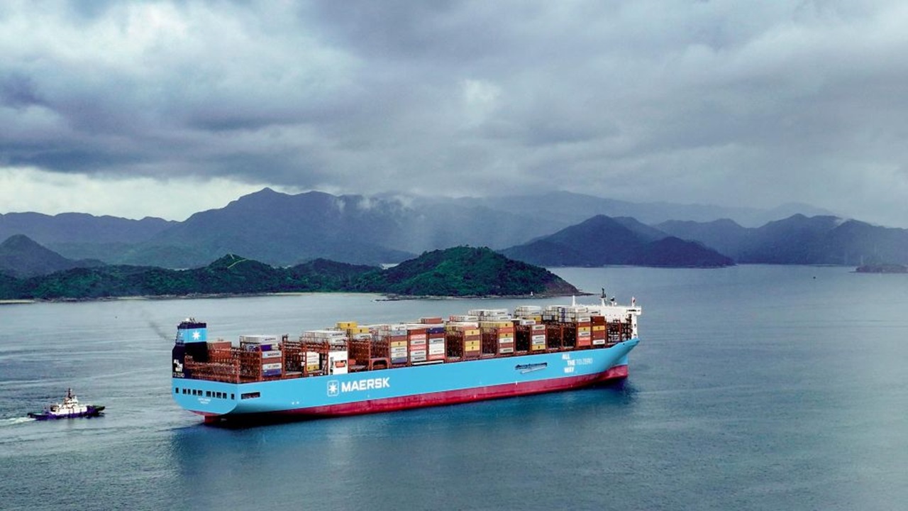 Maersk container ship carrying stacked cargo containers sailing on calm water near a mountainous coastline under cloudy skies.