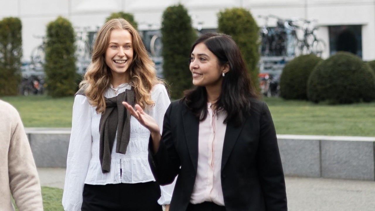 Two colleagues walking side by side outdoors, engaged in conversation, with office buildings, greenery, and bicycles in the background.