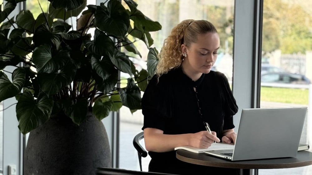 Person seated at a small table using a laptop and writing in a notebook, with a large indoor plant and office windows in the background.