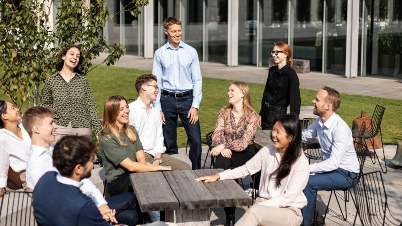 Group of people seated and standing around outdoor tables, engaged in a discussion in an office courtyard with a modern glass building in the background.