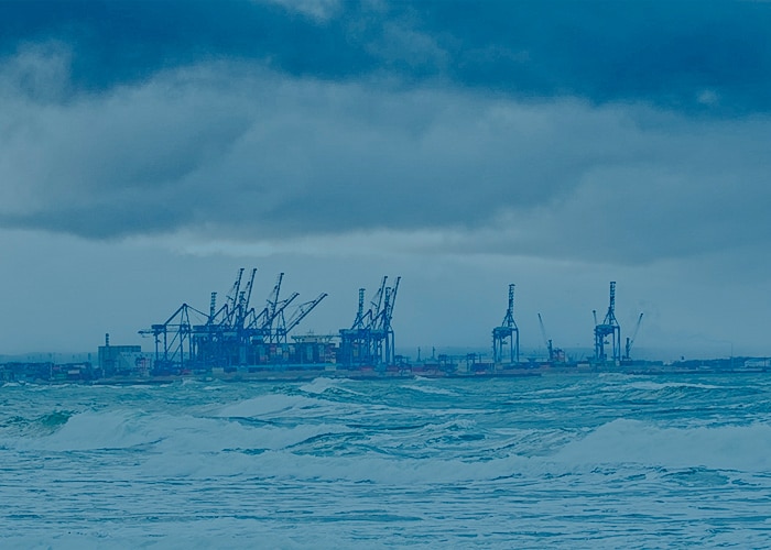 Stormy ocean waves with a view of a port featuring multiple container cranes under cloudy skies