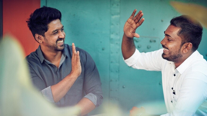 Two men with dark hair and brown skin smile and high five with a blue shipping container in the background. 