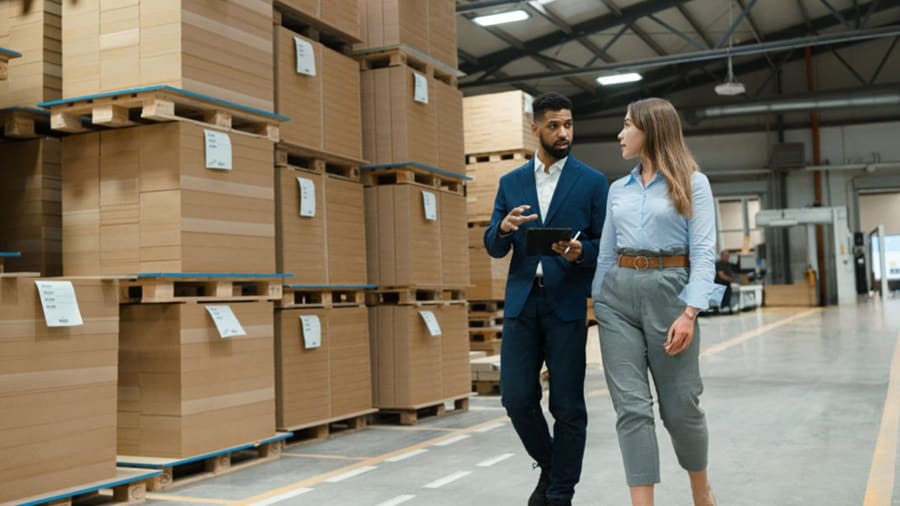 Two supply chain professionals walking through a warehouse with stacked cardboard pallets, discussing logistics operations and inventory strategy