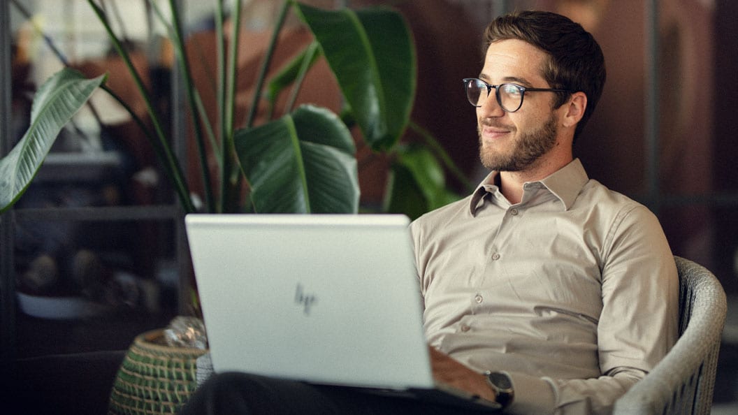 A man wearing glasses sits on a chair, working on a laptop.
