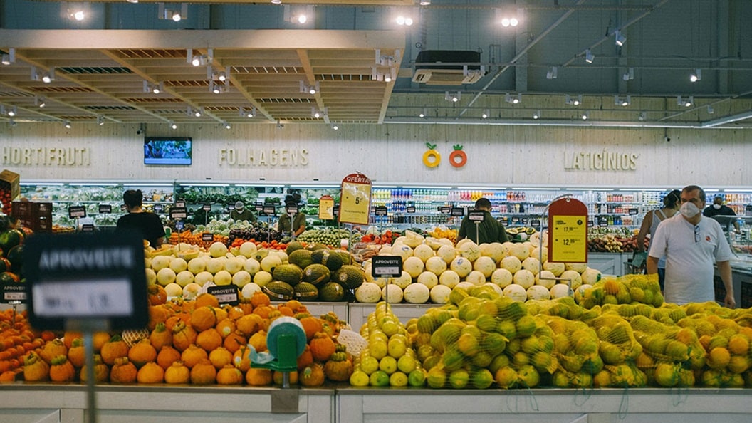 Wide view of a grocery store produce aisle with assorted fruits and vegetables and shoppers in the background
