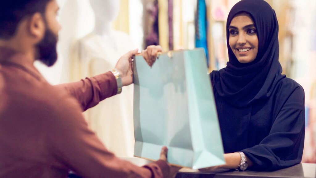 A woman wearing a hijab gives a shopping bag to a man, showcasing a moment of assistance and interaction.