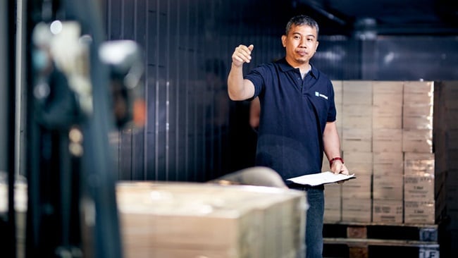 Person in a high-visibility vest using a manual pallet jack to move a pallet loaded with boxes