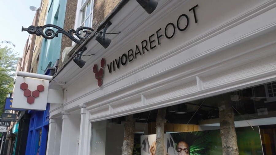 A woman in a stylish outfit is shopping, holding a sneaker and smiling as she examines it through a store window, with reflections adding depth to the scene.