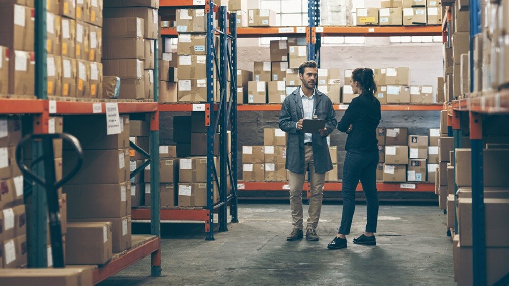 A male and a female conversing in a warehouse full of shipments