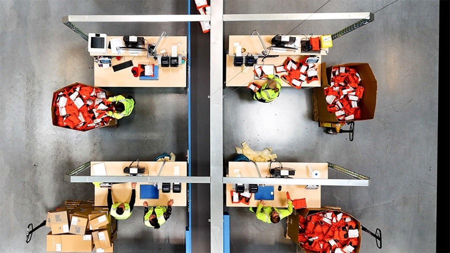 Top-down view of a logistics facility where workers in high-visibility vests process and sort e-commerce packages at organized workstations.