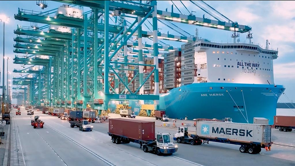 Large Maersk container ship docked at a busy port terminal, surrounded by cranes and cargo trucks.