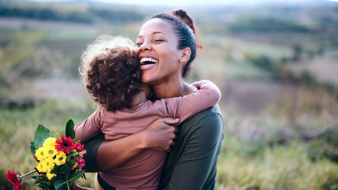 A woman and her child embrace warmly in a sunlit field, surrounded by tall grass and wildflowers.
