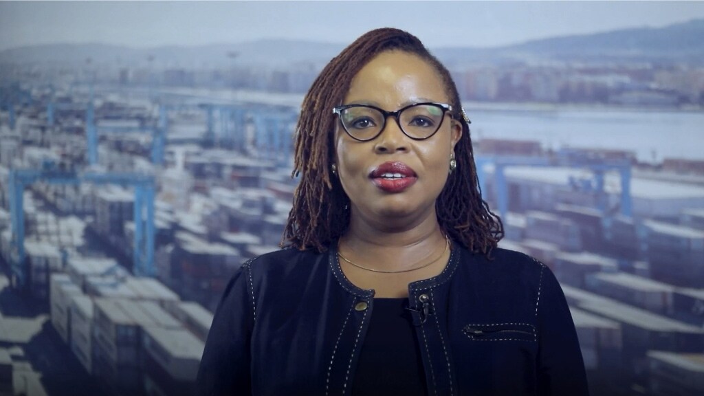 A woman wearing glasses stands in front of a large container ship, showcasing her interest in maritime activities.
