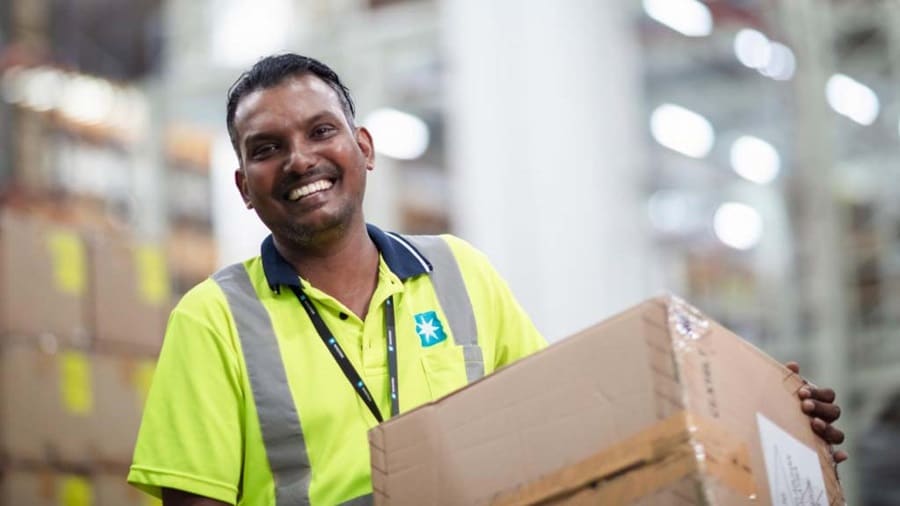 A cheerful man stands in a warehouse, holding a box, with storage shelves visible in the background.
