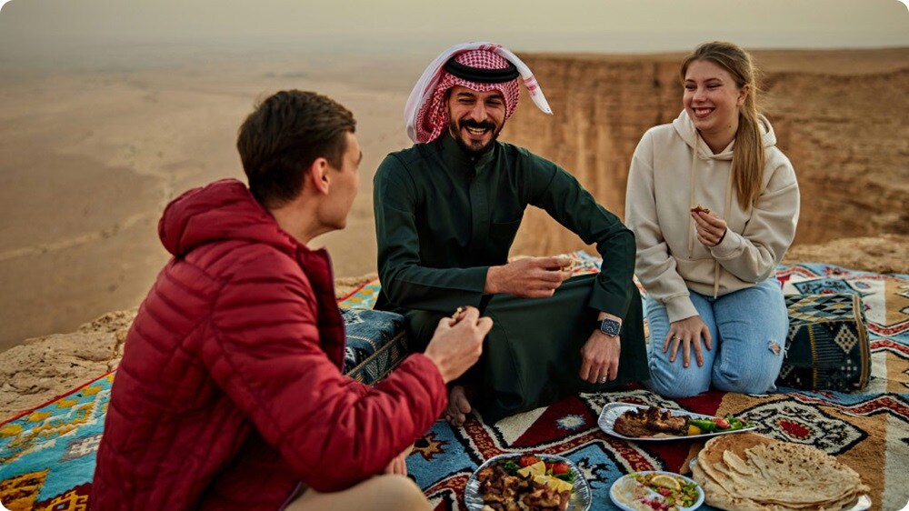 Three friends enjoying a traditional meal on rugs at a desert cliffside picnic.