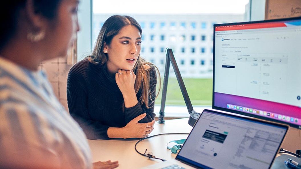 Two professionals are discussing shipping schedules on a computer screen in a modern office.
