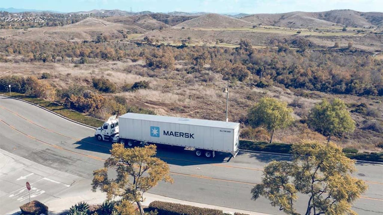 A Maersk container truck is driving on a road surrounded by trees and hills.