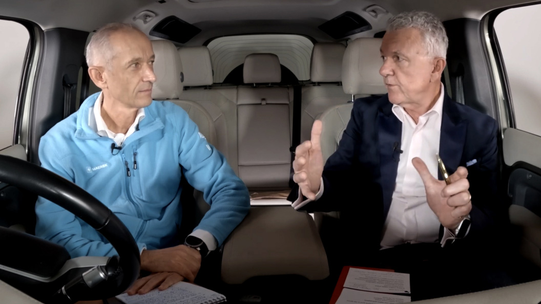 Two men sitting in the driver's seat of a car, both focused on the road ahead.