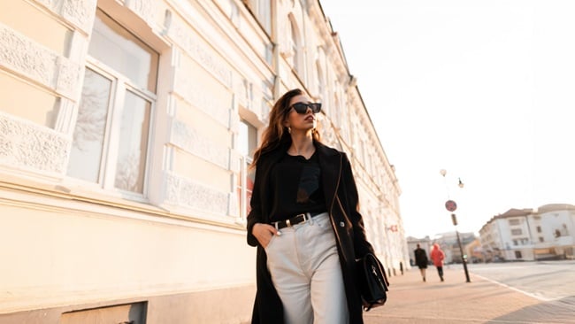 A woman in a black coat and white pants walks down a city street.