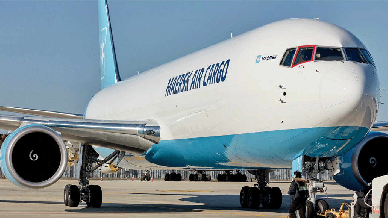 A Maersk Air Cargo plane is parked on the tarmac as a ground crew member works near the nose wheel.