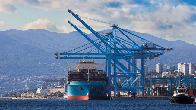 Large cargo ship docked at a port with blue container cranes, mountains in the background, and a partly cloudy sky.