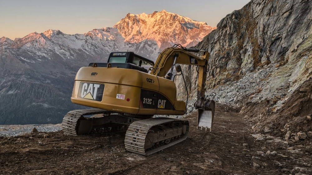 An industrial excavator parked on a mountainside, surrounded by rocky terrain and greenery.