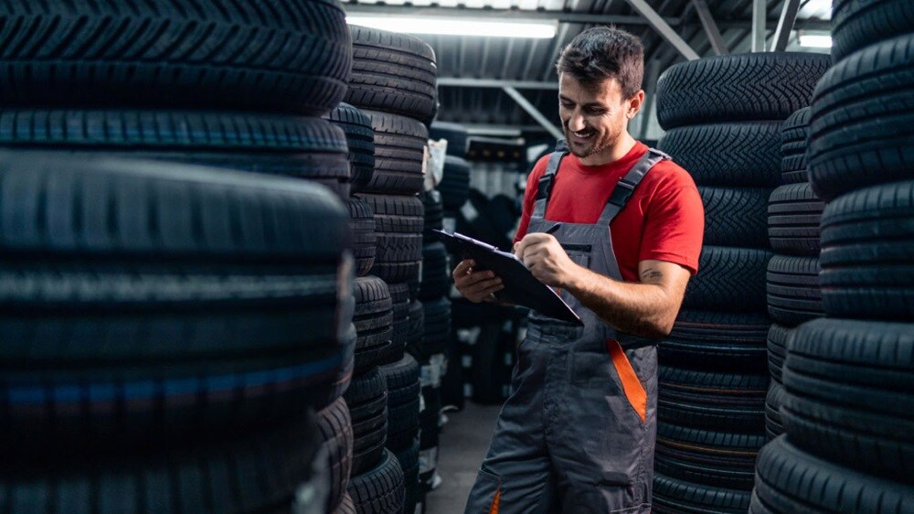 A man in an auto parts store holding a clipboard and reviewing inventory among shelves filled with car parts.