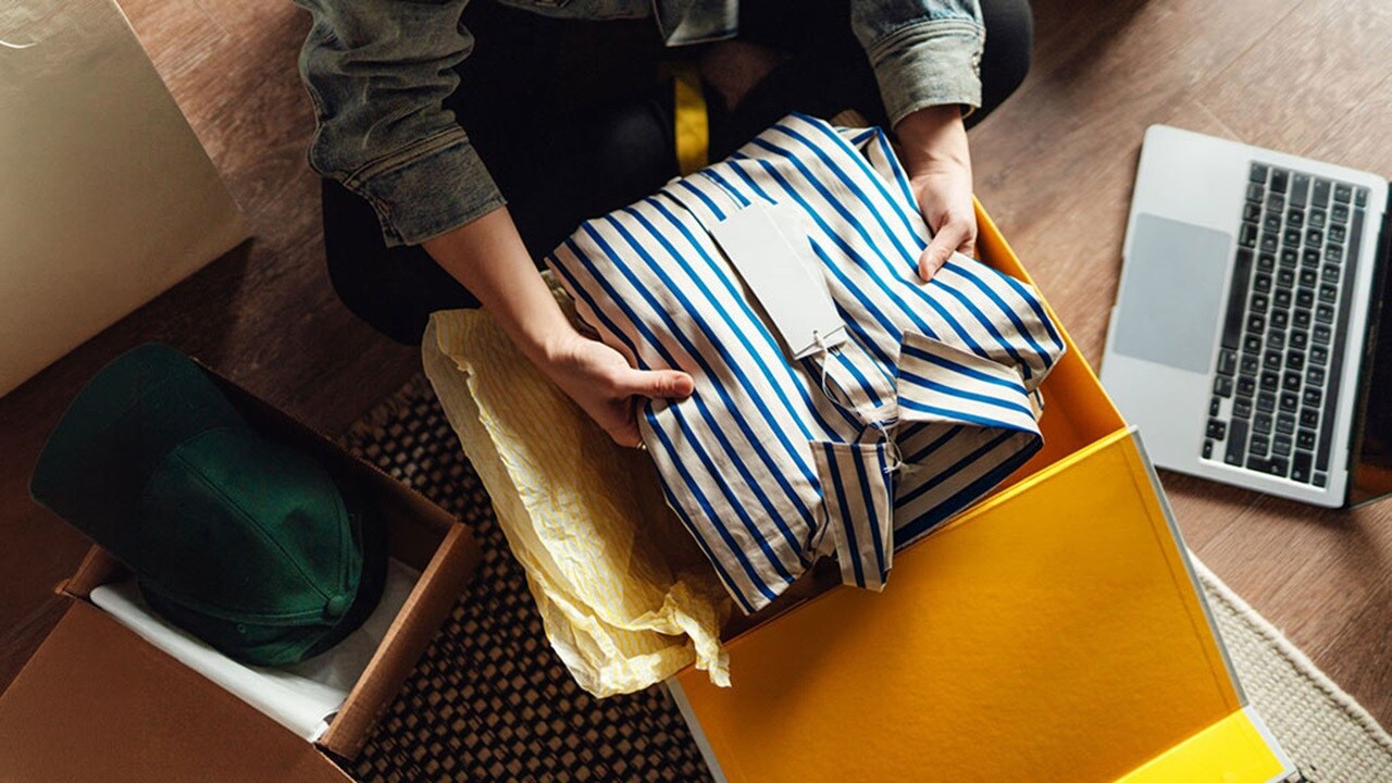 A woman sits on the floor with a laptop beside a box of clothes, focused on her work.