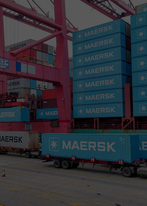 Maersk container being loaded onto a truck at a port terminal using a gantry crane, surrounded by stacked shipping containers